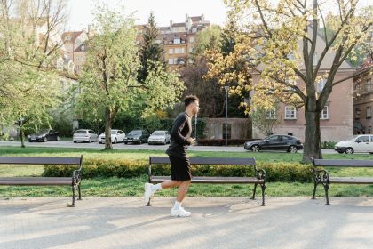 Homem corre em um parque arborizado, entre bancos e áreas urbanas, durante uma atividade física ao ar livre.