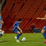 Jogadores disputam uma partida de futebol à noite, sob forte iluminação e diante de arquibancadas cheias.