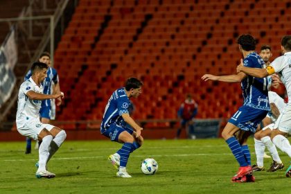 Jogadores disputam uma partida de futebol à noite, sob forte iluminação e diante de arquibancadas cheias.