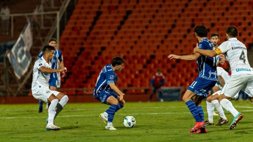 Jogadores disputam uma partida de futebol à noite, sob forte iluminação e diante de arquibancadas cheias.
