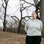 Mulher com corpo gordo, usando roupa esportiva, faz exercício em um parque durante o dia.