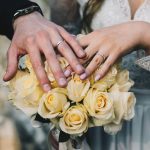 Mãos de um casal exibindo alianças sobre um buquê de rosas amarelas.