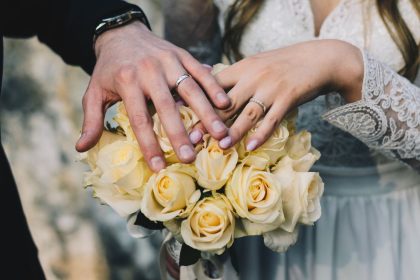 Mãos de um casal exibindo alianças sobre um buquê de rosas amarelas.