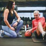 Homem idoso realiza exercícios em uma academia com orientação de um treinador.
