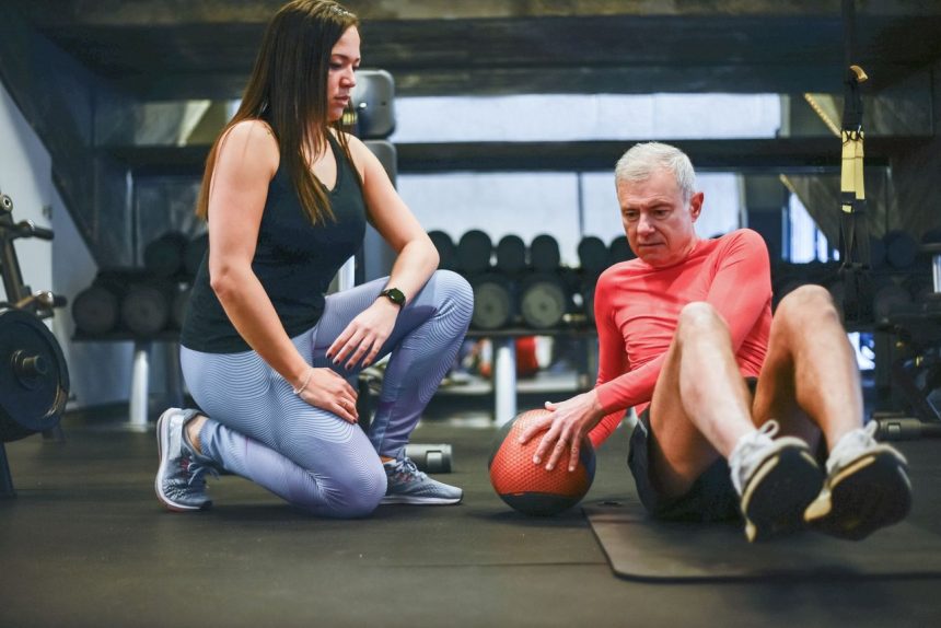 Homem idoso realiza exercícios em uma academia com orientação de um treinador.