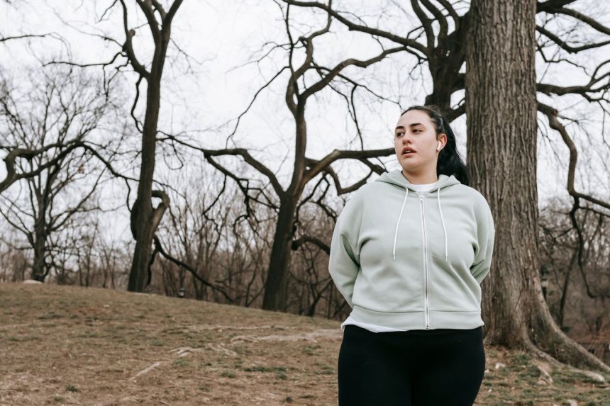 Mulher com corpo gordo, usando roupa esportiva, faz exercício em um parque durante o dia.
